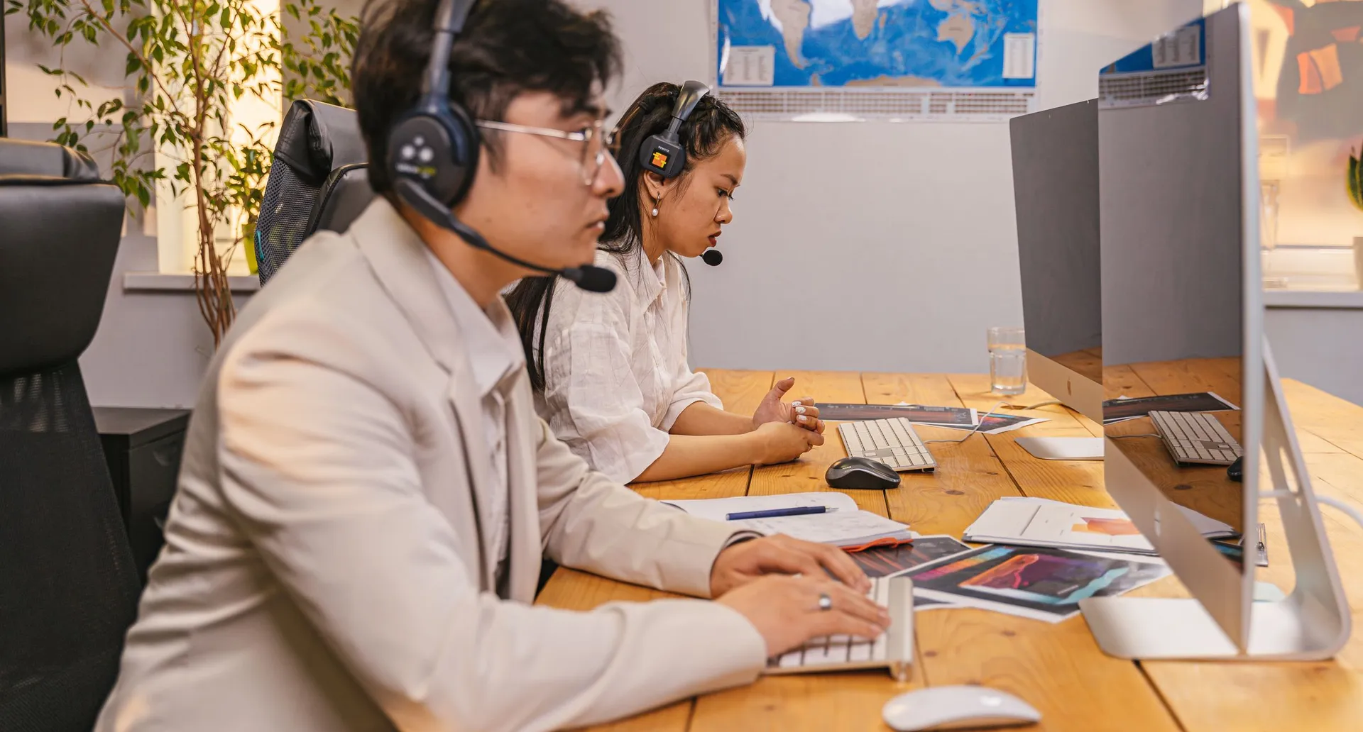 Two people working side by side at a shared desk, both in telephone headsets, monitors and keyboards in front of them.